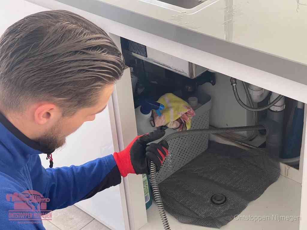 Een monteur voert een flexibele ontstoppingsveer in onder het keukenkastje, langs de sifon, om de gootsteen te ontstoppen in Nijmegen