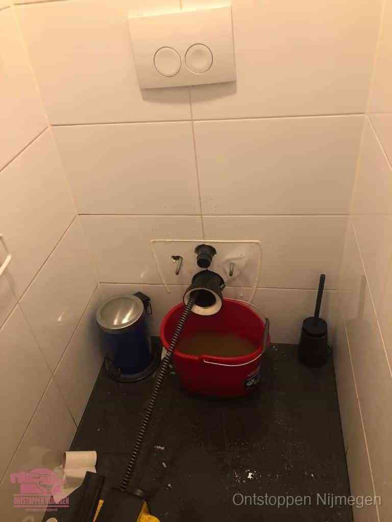 Een ontstopte wand-wc met verwijderde toiletpot waar een ontstoppingsveer in de wanduitgang verdwijnt, het achterhalen waar de verstopping zit in Nijmegen