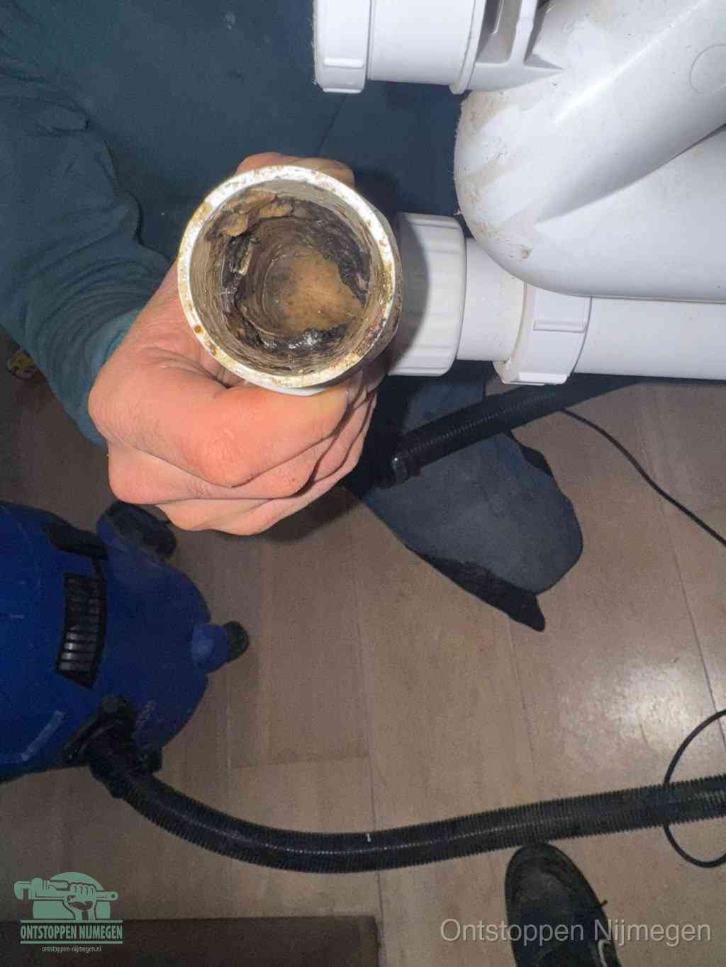 Een hand houdt een losgekoppeld PVC-afvoerbuisstuk vast dat van binnen volledig is dichtgeslibd met vet, vuil en aanslag, de meest voorkomende oorzaak van een verstopte afvoer in Nijmegen